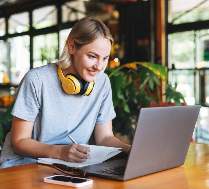 Young smiling blonde woman freelancer in with yellow headphones working on laptop on table at cafe